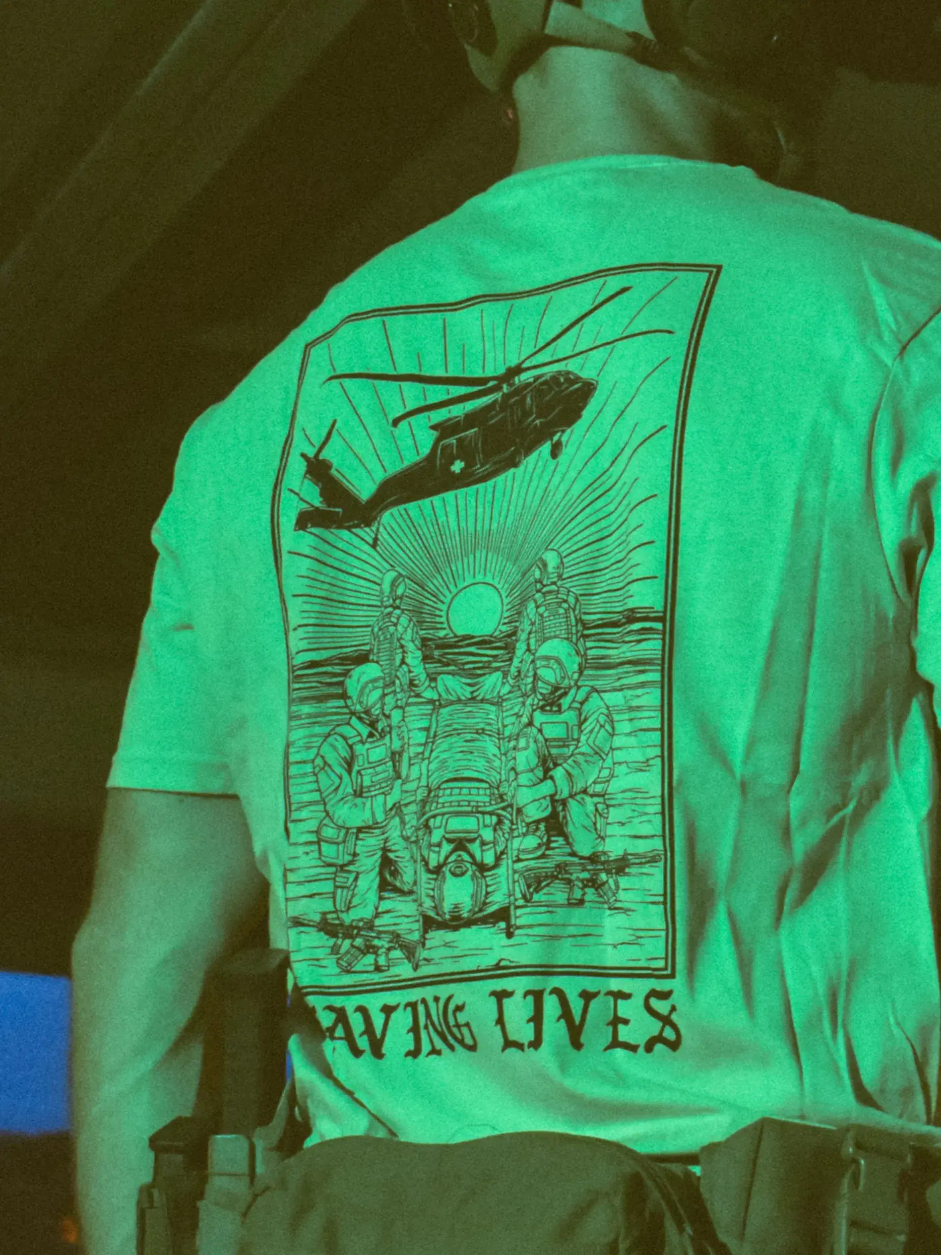 Cotton T-shirt with Saving Lives artwork showing soldiers in action, holding a stretcher during a tense medevac moment with a helicopter descending nearby.